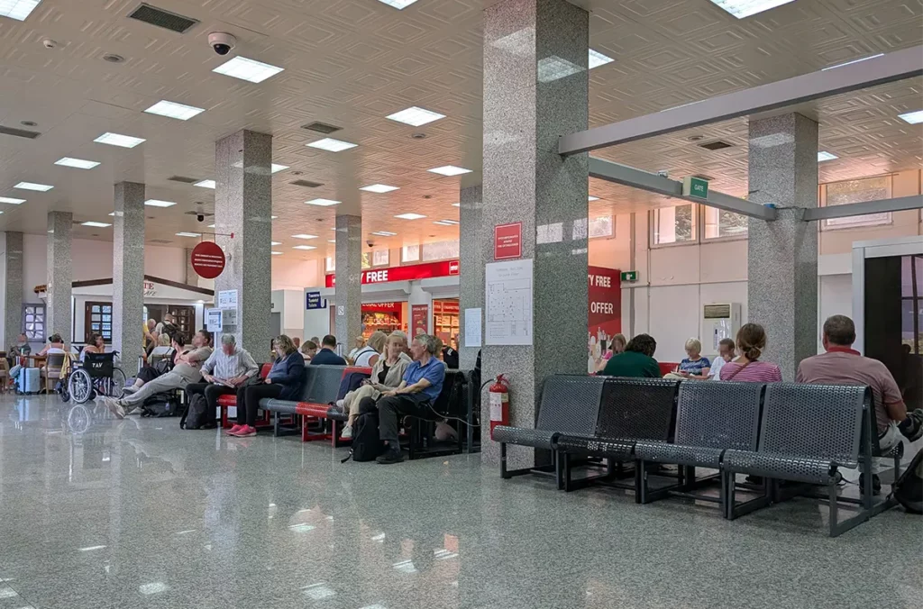 A small departures area at an airport. People are sat on seats. There's a small Duty Free shop and a little bar.