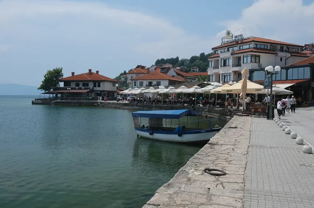 Lakeside restaurants with tables and chairs on a promenade. A little boat is moored on the quay.