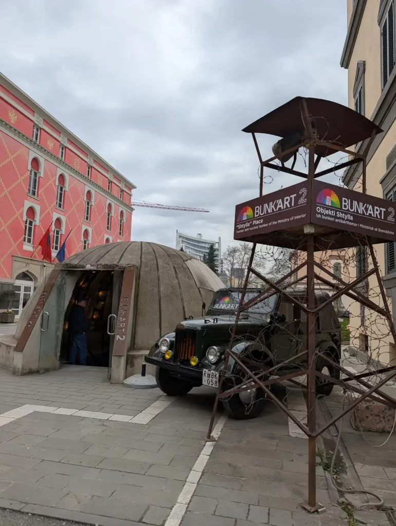 The entrance to Bunk'Art 2 in Tirana city centre. The actual entrance is under a replica concrete bunker dome. There's a jeep and a replica surveillance tower to the side of the entrance. 
