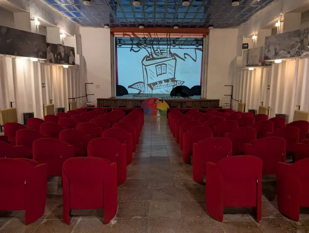The auditorium at Bunk'Art 1. Rows of red seats slope towards a stage at the front of a large, double height room.