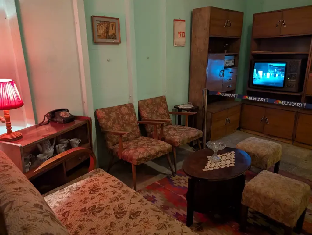 A recreation of an Albanian family's living room during the Communist regime. There are cosy chairs and a table. A TV in the corner is showing a political broadcast.