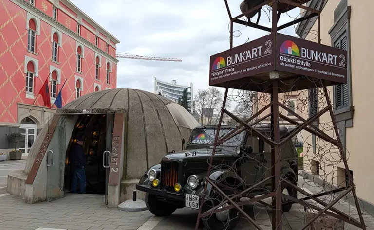 The entrance to Bunk'Art 2 in Tirana city centre. A concrete dome covers the entrance to the bunker museum. There's a military jeep and a surveillance tower to the side.