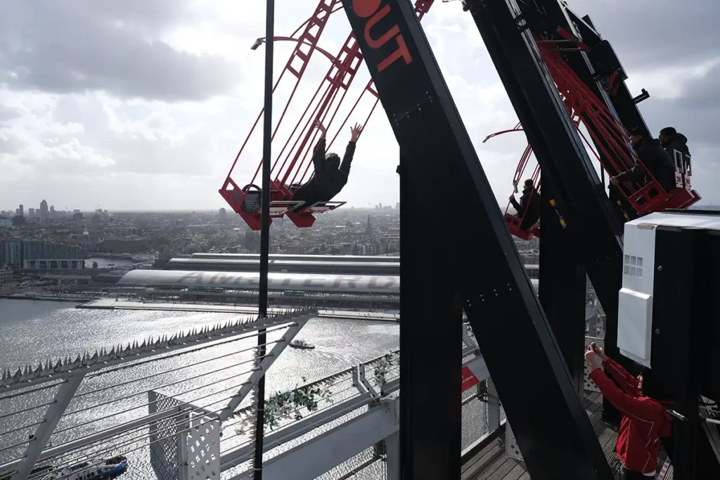 A swing swinging off the top of a tall building. A man is on the swing with his hands in the air.
