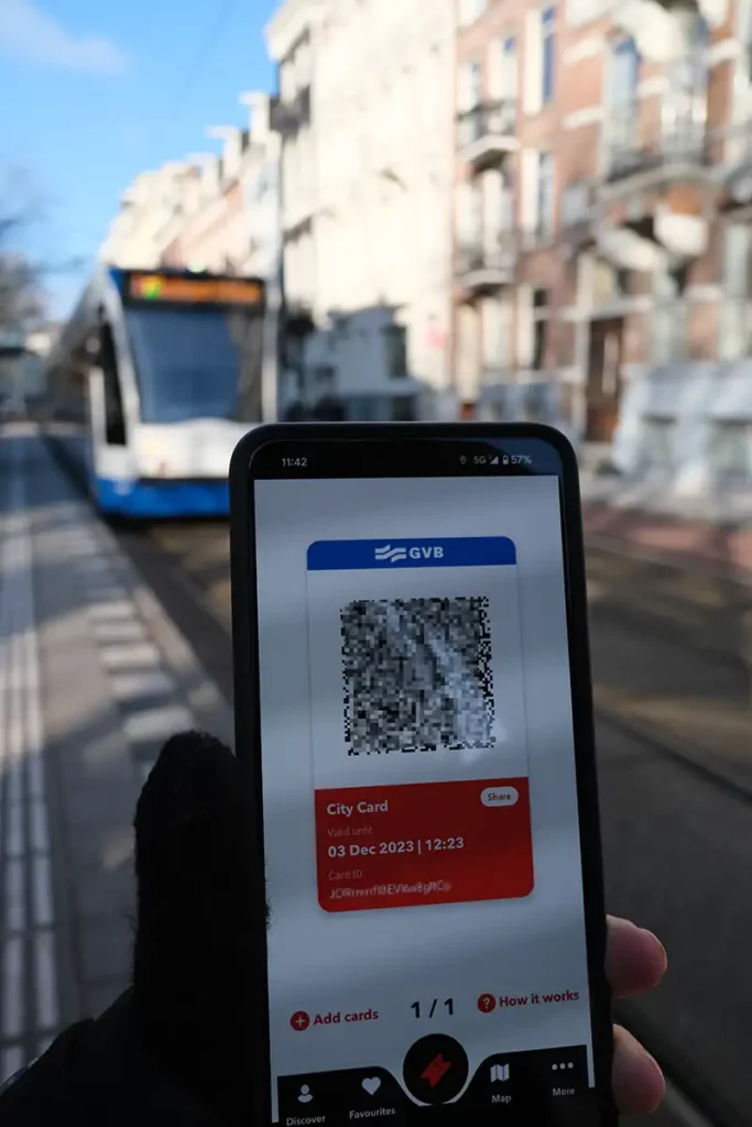 A gloved hand holding a phone. An I amsterdam digital city card is on the screen, with a QR code. A blue and white tram is approaching.