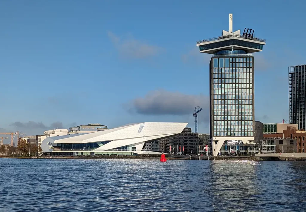 A modern, angular white museum building and a tall building with an observation deck on top, beside the river IJ in Amsterdam.