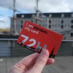 A hand holding a red I amsterdam city card 72 hours in front of an old building and sailing ship at the Maritime Museum, Amsterdam