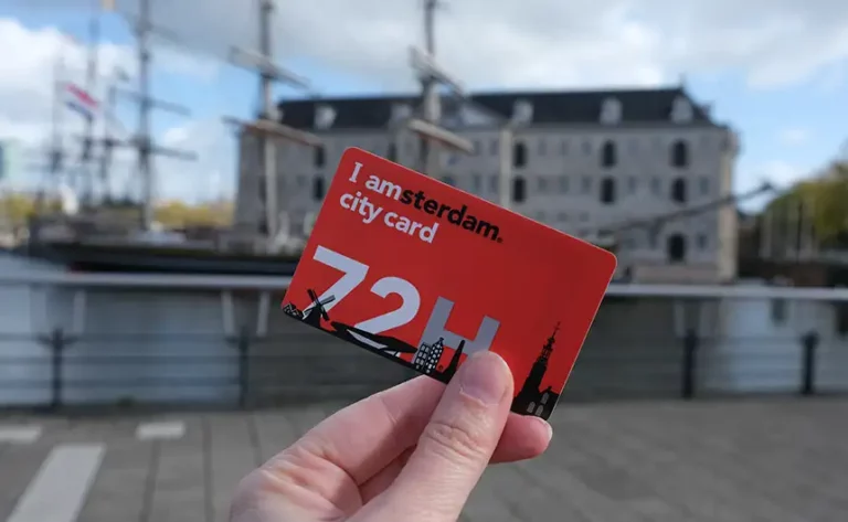 A hand holding a red I amsterdam city card 72 hours in front of an old building and sailing ship at the Maritime Museum, Amsterdam