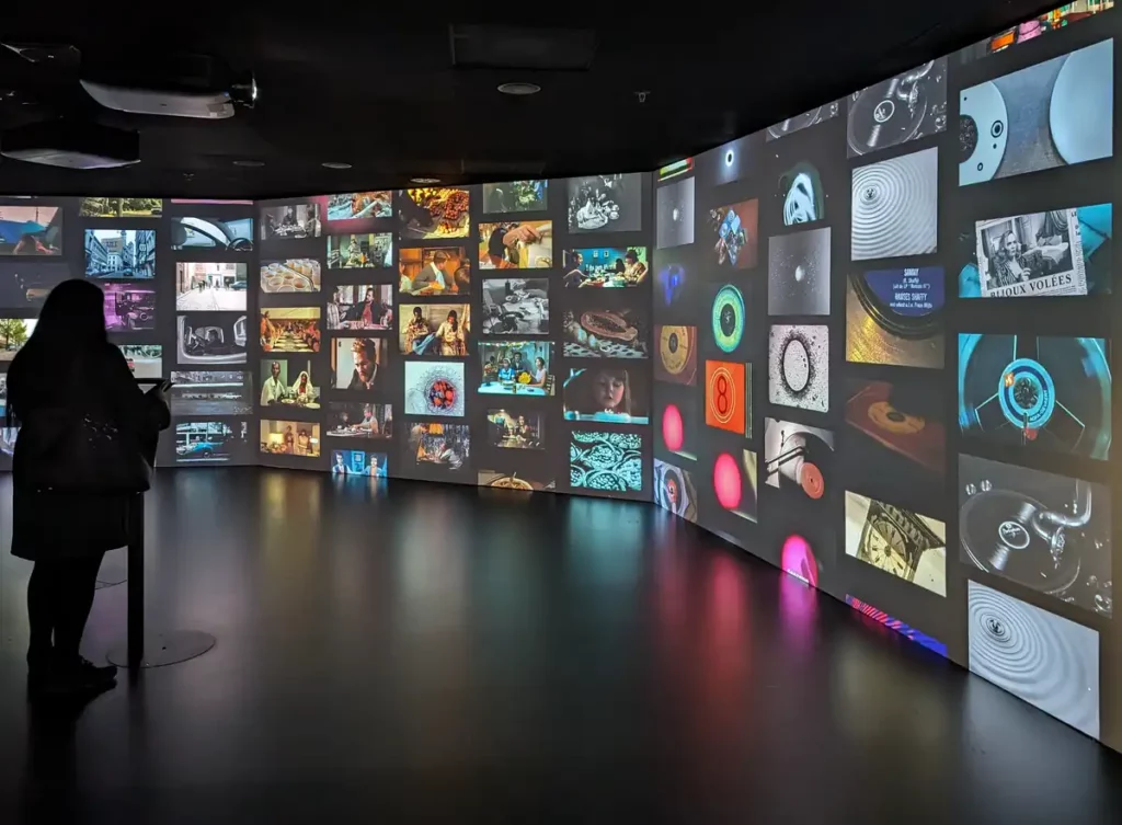 A wall in a black room, with lots of little screens all showing a different video. A woman is standing at a control screen, deciding what to watch next.