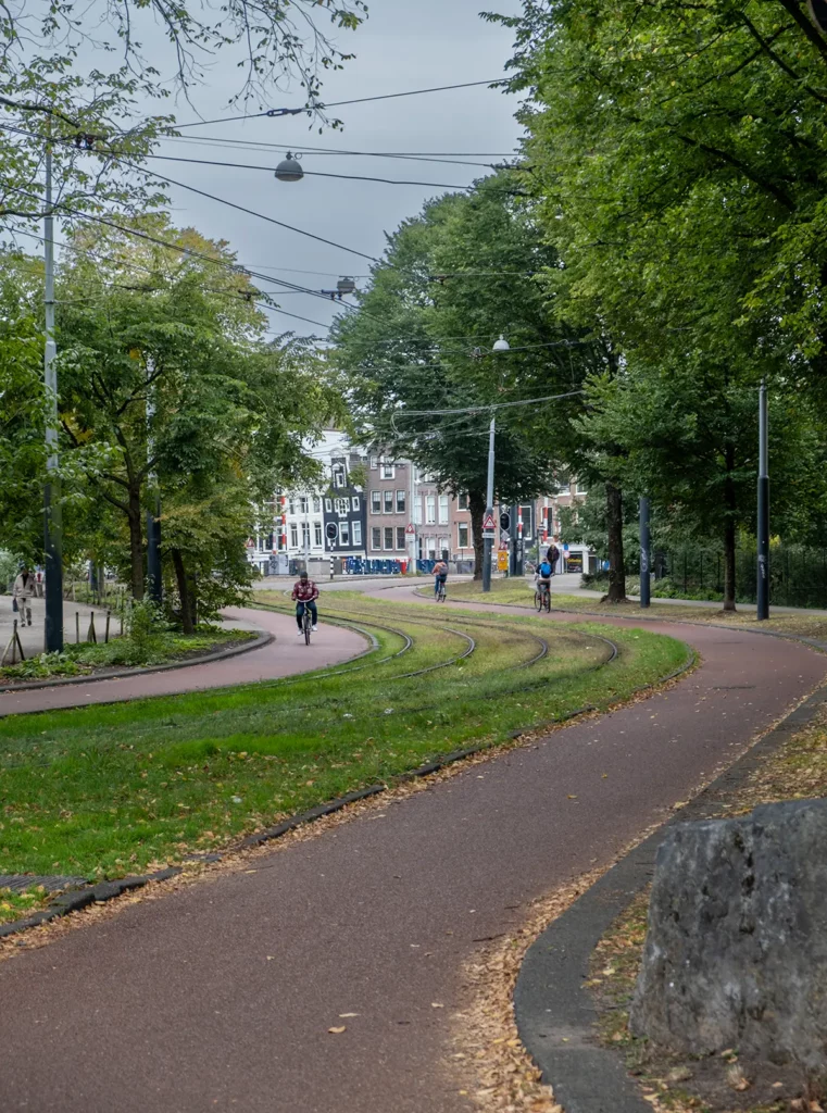 A tram track curving through a park with trees. There are cyclists and typically Dutch canal houses in the background.