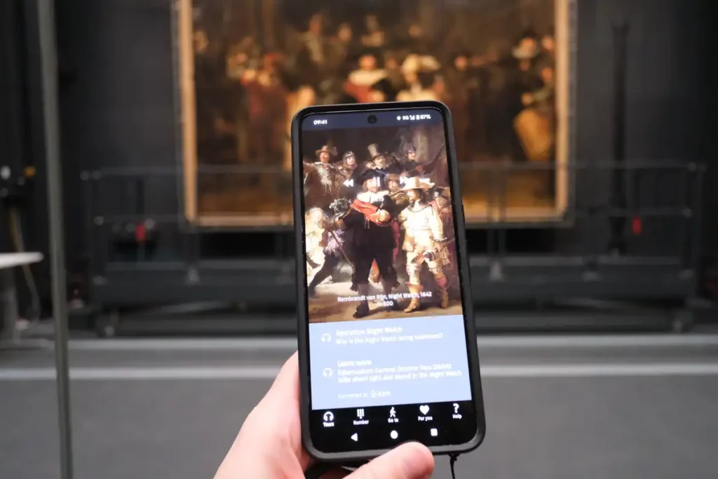 A hand holding a mobile phone. On the screen is information about Rembrandt's The Night Watch. The real The Night Watch painting is in the background. 