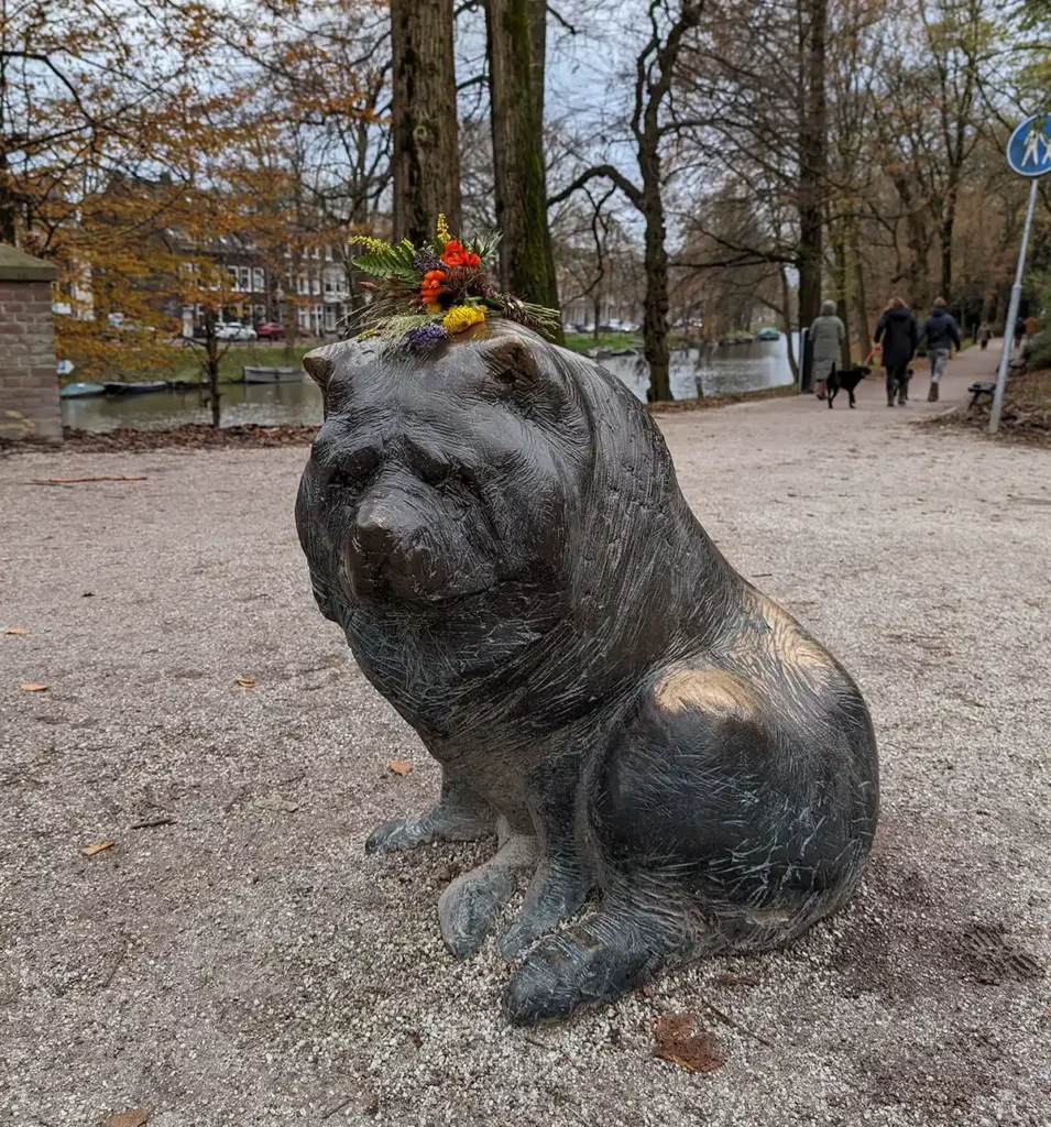 A statue of a dog in a park. People have left flowers on his head.