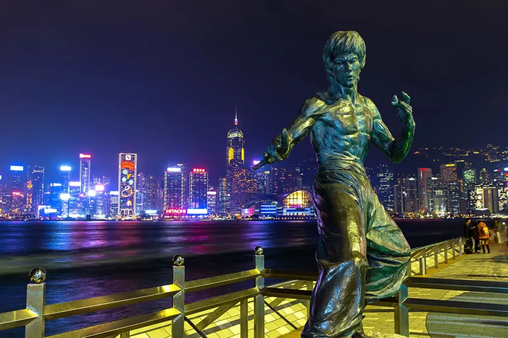A bronze statue of Bruce Lee on the Hong Kong harbour waterfront, with brightly lit skyscrapers in the background.