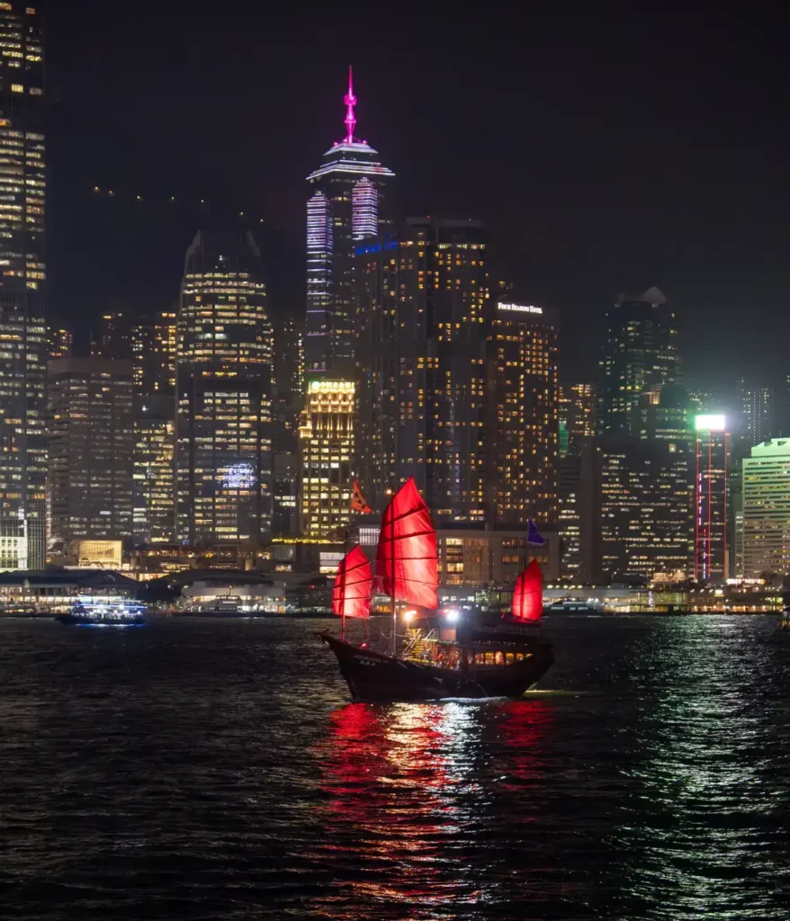 A night scene of a traditional sailing ship with red sails crossing Hong Kong's Victoria Harbour in front of skyscrapers