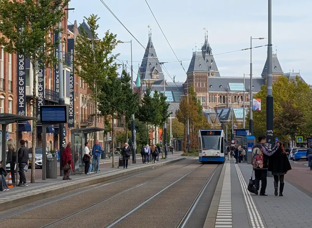 A tram arriving at the Museumplein tram stop. There is a large red brick building in the distance, the Rijksmuseum.