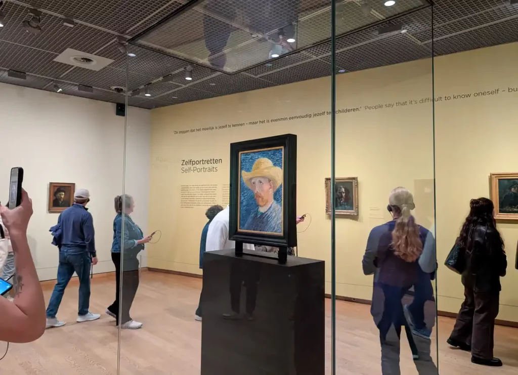 People in a gallery. One of Van Gogh's self portraits is displayed in the middle of the room.