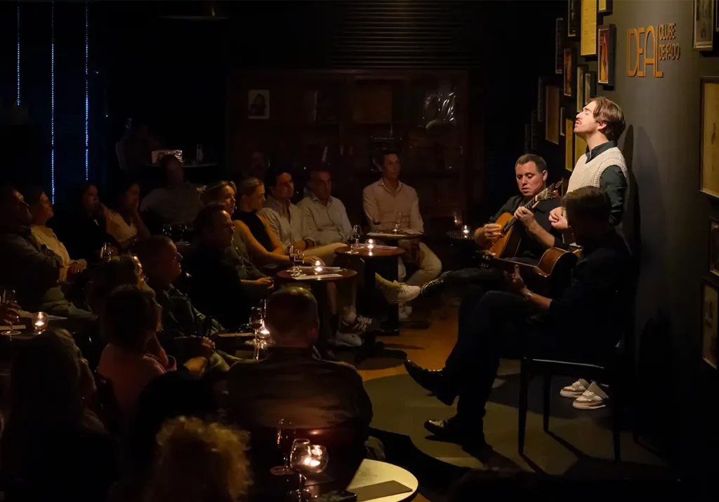 An audience sat around small stage in a softly-lit room, with two guitarists and a singer performing fado at Ideal Club de Fado in Porto