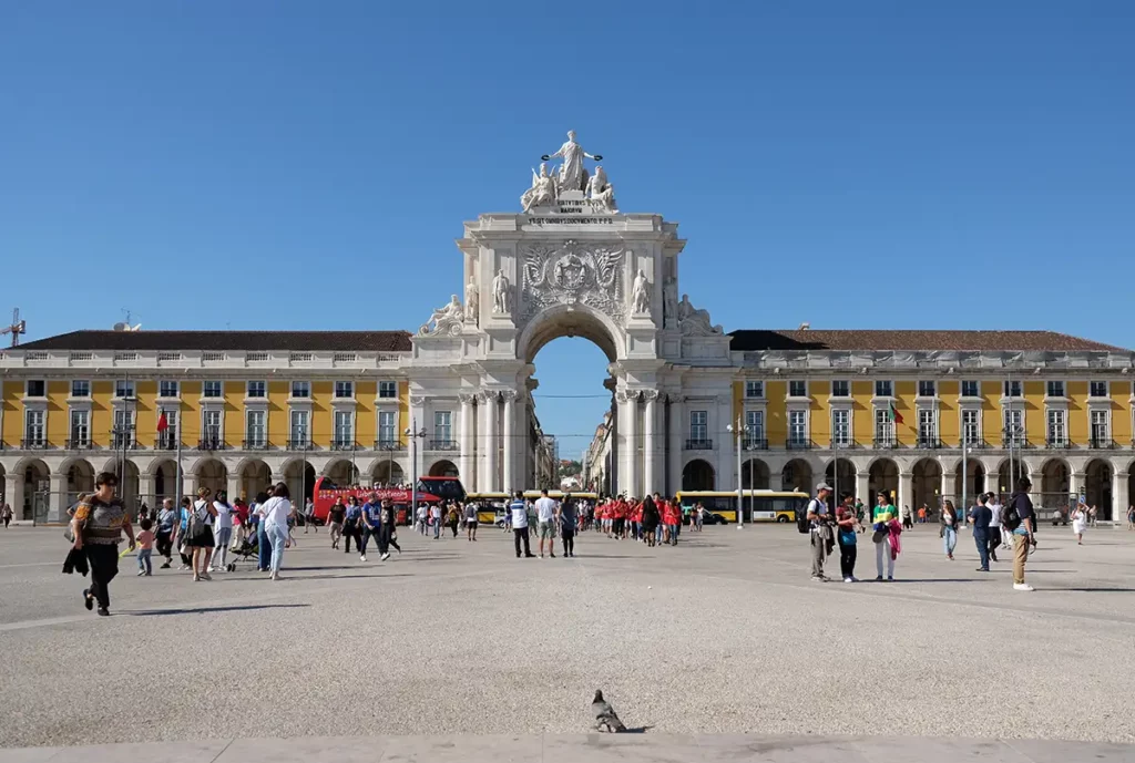 Praça do Comércio in Lisbon, Portugal. A huge, ornate white arch is flanked by a grand yellow building in front of a huge square